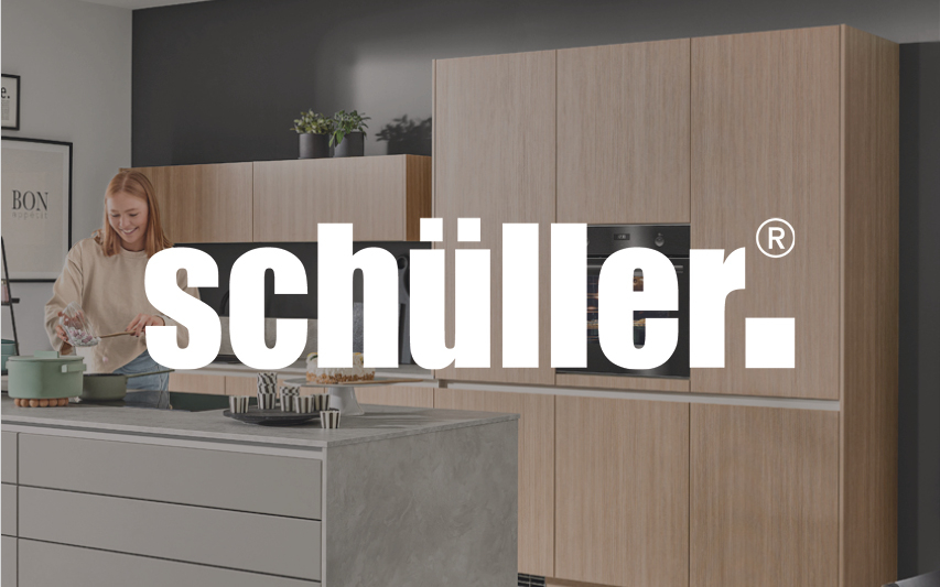 Moderne Schüller-Küche mit Holzfronten und Kochinsel in Betonoptik, mit einer Frau beim Kochen und großem Schüller-Logo im Vordergrund.