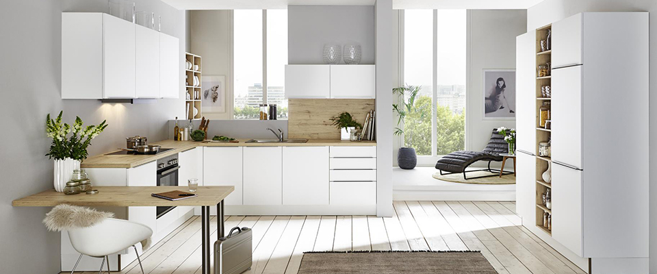 Helle, moderne Küche in L-Form mit weißen Fronten, Holzarbeitsplatten und integrierten Regalen; große Fenster lassen viel Tageslicht herein, im Hintergrund eine gemütliche Sitzecke mit Liegestuhl.