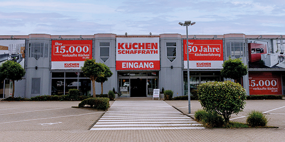 Das Bild zeigt den Eingangsbereich eines K&uuml;chenfachmarkts mit dem Schriftzug &bdquo;K&uuml;chen Schaffrath&ldquo; &uuml;ber dem Haupteingang. An der Fassade sind mehrere gro&szlig;formatige Tafeln mit Hinweisen zu Unternehmenszahlen und Jubil&auml;en angebracht. Ein markierter Fu&szlig;g&auml;nger&uuml;berweg f&uuml;hrt &uuml;ber den Vorplatz direkt zum Eingang. Das Geb&auml;ude ist von bepflanzten Gr&uuml;nfl&auml;chen und einem Parkplatz umgeben.