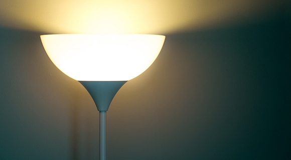 Leuchtende Stehlampe mit nach oben gerichtetem Lichtkegel vor dunkler Wand.