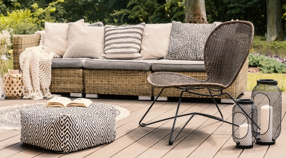 Gartenlounge aus Geflecht mit Sitz- und R&uuml;ckenkissen, davor ein Pouf und Laternen auf einer Holzterrasse.