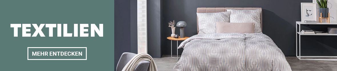 Werbebanner mit grünem Hintergrund links, weißer Aufschrift „TEXTILIEN“ und Button „MEHR ENTDECKEN“. Rechts ein stilvoll eingerichtetes Schlafzimmer mit gemusterter Bettwäsche in Grau-Beige auf einem gepolsterten Bett, flankiert von Nachttisch und Konsole mit Deko. Kleine Blümchen-Symbole lockern das Design auf.