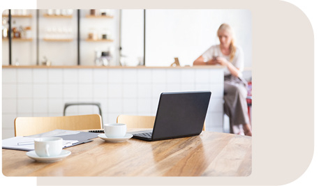 Offener Bürobereich mit einem Holztisch, auf dem ein Laptop und eine Kaffeetasse stehen. Im Hintergrund sitzt eine Frau entspannt an einer Theke mit Fliesenrückwand und Regalen mit dekorativen Gegenständen. Der Raum wirkt hell und einladend, mit einer Mischung aus Arbeits- und Pausenbereich