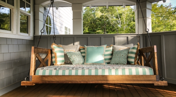 Hollywoodschaukel aus Holz mit gemusterten Kissen auf einer Veranda mit Blick ins Gr&uuml;ne.