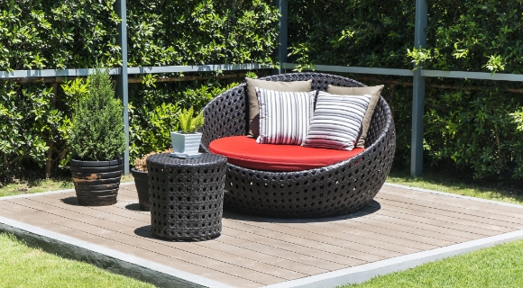 Runde Outdoor-Liege aus Polyrattan auf Holzdeck mit roter Sitzauflage und gestreiften Kissen, daneben kleiner Beistelltisch und Pflanzt&ouml;pfe.