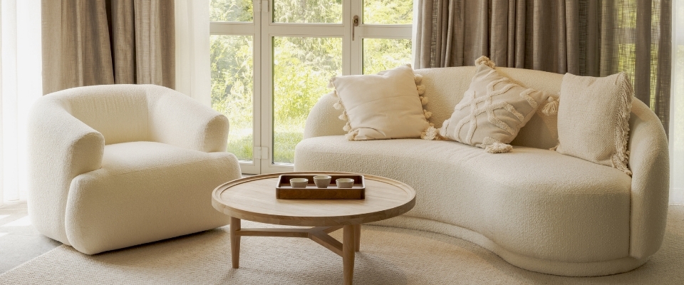 Helles Wohnzimmer mit cremefarbenem Sofa, Sessel und rundem Holztisch vor gro&szlig;en Fenstern mit Blick ins Gr&uuml;ne.