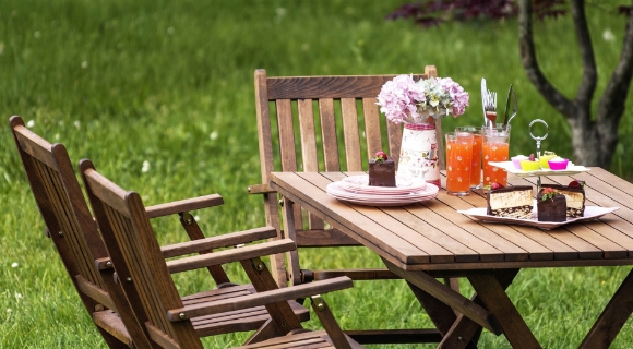 Holzgartentisch mit zwei Klappst&uuml;hlen, gedecktem Geschirr, Getr&auml;nken und Kuchen auf einer Rasenfl&auml;che.
