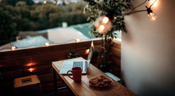 Ein gem&uuml;tlicher Balkon mit einem Laptop auf einem Holztisch, einer Tasse Kaffee und einem Teller mit Keksen. Lichterketten h&auml;ngen im Hintergrund.