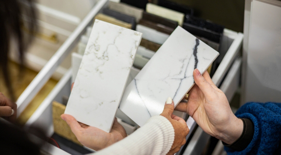 Zwei Hände halten nebeneinander zwei Musterplatten mit Marmoroptik über einer geöffneten Schublade mit weiteren Materialmustern.