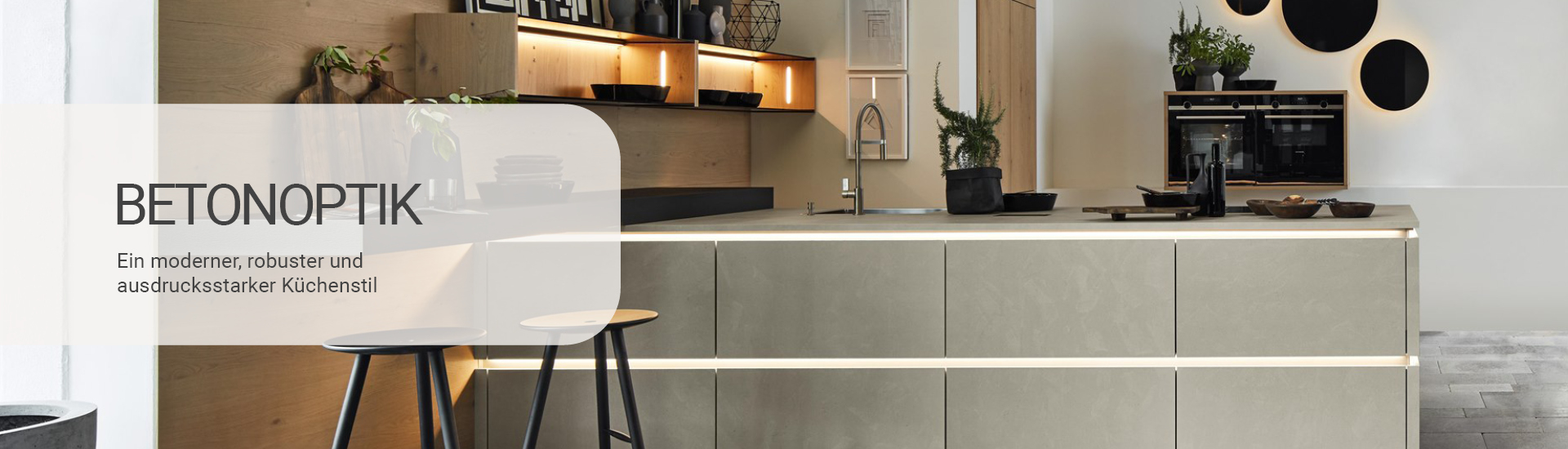 Küche in Betonoptik mit moderner, robuster Gestaltung. Links im Bild ein Textfeld mit der Aufschrift Betonoptik – Ein moderner, robuster und ausdrucksstarker Küchenstil'. Die Küche verfügt über eine große Insel mit Betonoptik und integrierter Beleuchtung. Die Rückwand ist mit offenen Regalen und Dekorationen versehen, und runde schwarze Wanddekorationen setzen einen zusätzlichen Akzent