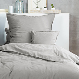 Hellgraue Bettwäsche mit glatter Oberfläche auf aufgeschütteltem Bett, kombiniert mit Deko-Elementen auf weißem Regal – schlichte und moderne Schlafzimmereinrichtung.