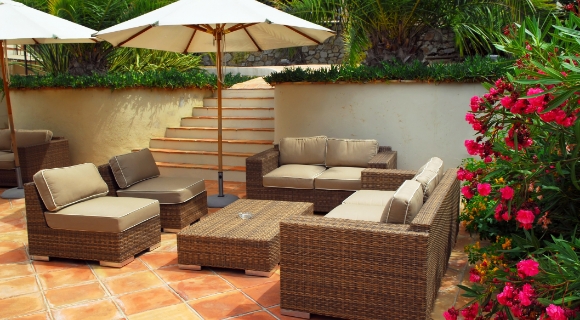 Outdoor-Lounge aus Rattanmöbeln mit Polstern, rechteckigem Tisch und angrenzenden Palmen und blühenden Sträuchern.