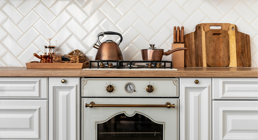 Landhausküche mit einem cremefarbenen Gasofen im Vintage-Stil und einer Arbeitsplatte aus Holz. Auf dem Herd stehen ein kupferfarbener Teekessel und ein Topf. Auf der Arbeitsfläche befinden sich außerdem Schneidebretter, ein Messerblock und weitere Küchenutensilien. Die Rückwand ist mit weißen Fliesen im Fischgrätenmuster verkleidet