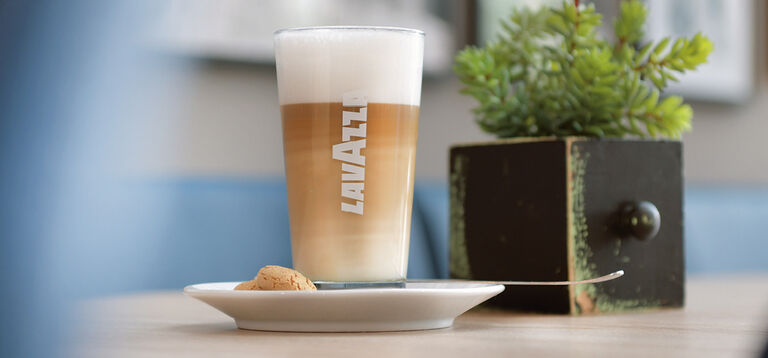 Das Bild zeigt ein Latte-Macchiato-Glas mit dem weißen Schriftzug „Lavazza“ auf der Vorderseite. Der Latte Macchiato ist in drei klaren Schichten erkennbar: unten brauner Espresso, in der Mitte milchiger Kaffee und oben cremiger Milchschaum. Das Glas steht auf einem weißen Unterteller, auf dem ein kleiner Keks liegt. Rechts vom Glas befindet sich ein moderner Holzwürfel mit grünen, künstlichen Pflanzen, die eine gemütliche Café-Atmosphäre schaffen. Im Hintergrund sind unscharf weitere Dekoelemente und ein heller Raum sichtbar, der die Szene warm und einladend wirken lässt.