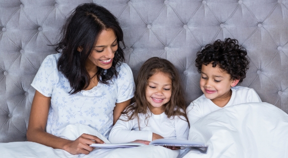 Mutter liest im Bett gemeinsam mit zwei Kindern ein Buch – gemütliche Familienszene vor gesteppter Kopfteilpolsterung in einem großzügigen Bett