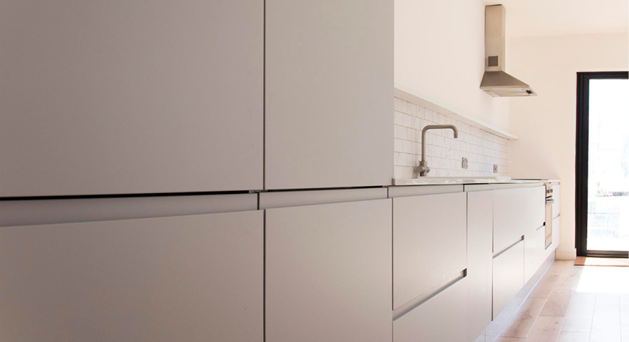 Minimalistische Küche mit glatten, grifflosen Fronten in neutralen Tönen. Die Arbeitsfläche ist schlicht gestaltet, mit einer einzelnen Armatur und einem weißen Fliesenspiegel im Hintergrund. Am Ende der Küche befindet sich ein großes Fenster, das viel Licht in den Raum bringt