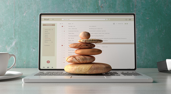 Laptop mit geöffnetem E-Mail-Postfach, davor ein aufgestapelter Turm aus Steinen als Symbol für Stabilität und Balance.
