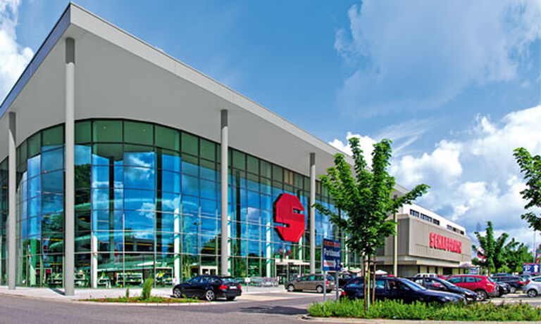 Das Bild zeigt ein modernes, großflächiges Gebäude mit einer Fassade aus Glas und Metall. Im Zentrum der Glasfront spiegelt sich der blaue Himmel mit weißen Wolken. Auf der linken Seite ist ein großes, rotes "S"-Logo deutlich zu erkennen, das die Marke Schaffrath repräsentiert. Rechts im Bild sieht man den Schriftzug "Schaffrath" auf einem zweiten Gebäudeteil. Vor dem Gebäude befinden sich mehrere parkende Autos sowie ein Bereich mit kleinen Bäumen und Gehwegen. Der Himmel ist klar und sonnig, was eine helle und einladende Atmosphäre erzeugt.