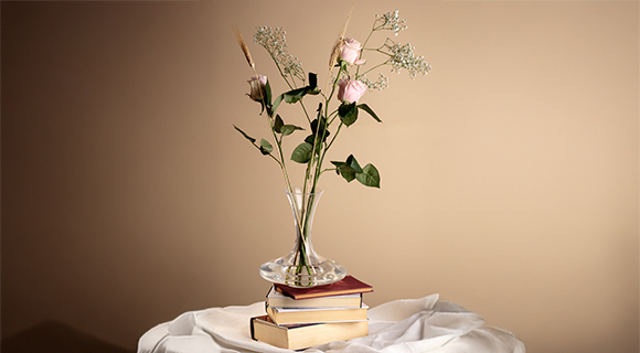 Eine transparente Glasvase mit zarten rosa Rosen und Zweigen steht auf einem Stapel Bücher. Die Szene ist schlicht arrangiert und wird von warmem Licht hervorgehoben.