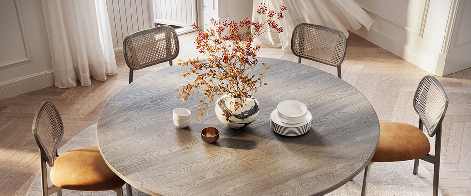 Runder Holztisch mit sechs gepolsterten Stühlen auf einem runden Teppich in einem hellen Raum mit Parkettboden und großen Fenstern; auf dem Tisch stehen Geschirr und eine Vase mit Zweigen.