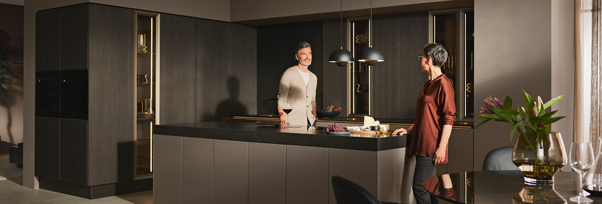 Zwei Personen in einer minimalistischen, in Brauntönen gehaltenen Küche, in die von rechts die Morgensonne durch ein Fenster scheint