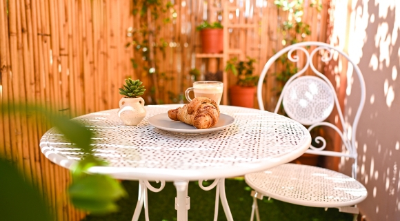 Ein kleiner Balkon mit einem wei&szlig;en Metalltisch, auf dem ein Croissant, eine Tasse Kaffee und eine kleine Pflanze stehen. Der Balkon ist von einem Bambuszaun und Pflanzen umgeben.