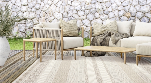 Gemütliche Terrasse mit hellen Loungemöbeln, Holztischen und beigem Outdoor-Teppich auf Holzdeck vor Natursteinmauer.