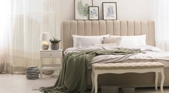 Hell eingerichtetes Schlafzimmer mit Boxspringbett und hohem, gepolstertem Kopfteil als Mittelpunkt im Raum, grüner Tagesdecke und dekorativen Wandbildern