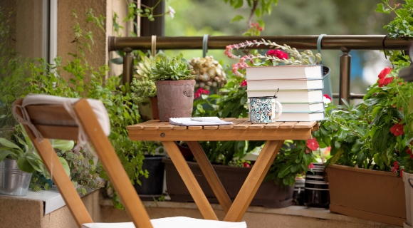 Ein Balkon mit einem Holzstuhl und einem Tisch, auf dem ein Stapel B&uuml;cher, eine Tasse mit einem L&ouml;ffel und ein Notizbuch liegen. Der Balkon ist von gr&uuml;nen Pflanzen und Blumen umgeben.