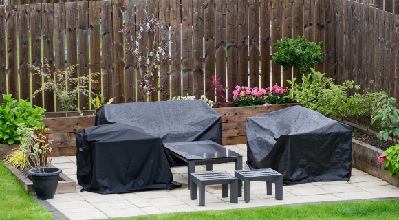 Terrasse im Garten mit abgedecktem Grill und abgedeckter Sitzgruppe, davor kleiner Tisch mit zwei Hockern, Hochbeete und Holzzaun im Hintergrund.
