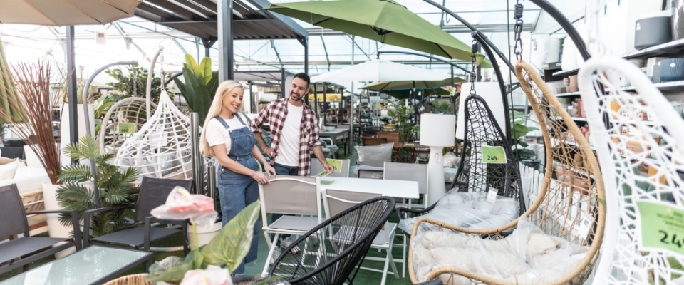 Zwei Personen in einem M&ouml;belmarkt zwischen Outdoor-M&ouml;beln, darunter H&auml;ngesessel, Tische und Sonnenschirme in einer gro&szlig;en Ausstellungsfl&auml;che.