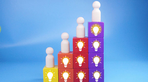 Holzfiguren stehen auf farbigen Würfeln mit Glühbirnensymbolen, die eine ansteigende Treppenform bilden.