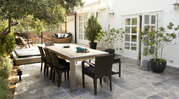 Terrasse mit gro&szlig;em Holztisch, vier St&uuml;hlen, Pflanzk&uuml;beln und wei&szlig;er Hausfassade.