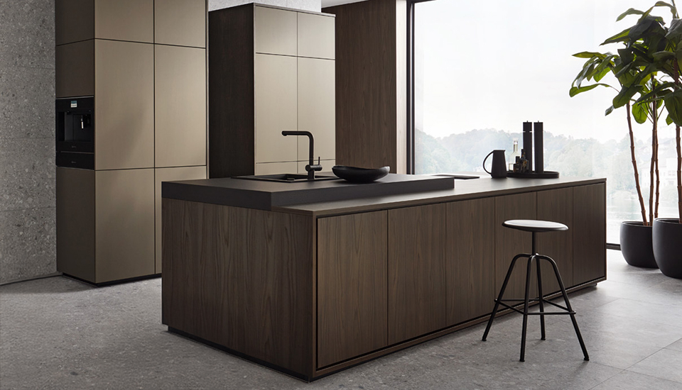 Moderne, minimalistisch gestaltete Küche mit einer großen Kochinsel im Vordergrund, die aus dunkel gemasertem Holz besteht und mit einer mattschwarzen Arbeitsplatte versehen ist. Auf der Insel befinden sich ein flacher Wasserhahn, eine eingelassene Spüle sowie dekorative Elemente wie ein flacher schwarzer Teller, eine Kanne und Gewürzbehälter. Rechts daneben steht ein schlichter schwarzer Hocker mit weißer Sitzfläche. Im Hintergrund ist eine hohe Schrankwand mit glatten, grifflosen Fronten in Beige zu sehen, in die ein Kaffeevollautomat integriert ist. Durch das bodentiefe Fenster fällt Tageslicht ein, und eine große grüne Zimmerpflanze in einem schwarzen Topf bringt etwas Natürlichkeit in den ansonsten puristischen Raum. Der Boden besteht aus grauen Fliesen, die den modernen Look unterstreichen.