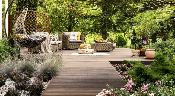 Holzterrasse im Garten mit H&auml;ngesessel aus Geflecht, Lounge-Sofa mit Kissen, Pouf und niedrigem Tisch, umgeben von Pflanzenbeeten.