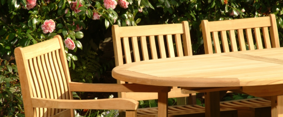 Runder Gartentisch aus Holz mit mehreren Holzst&uuml;hlen vor bl&uuml;henden Str&auml;uchern.