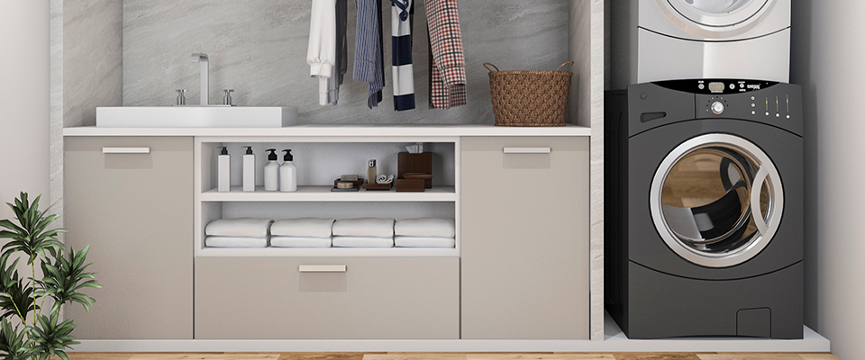 Moderner Hauswirtschaftsraum mit beige-weißen Einbauschränken, offenem Regal und integriertem Waschbereich. Links befindet sich ein rechteckiges Waschbecken mit Edelstahlarmatur auf einer weißen Arbeitsplatte. Darunter stehen mehrere Seifenspender und gefaltete Handtücher ordentlich in offenen Fächern. In der Mitte hängt Kleidung an einer Kleiderstange, darüber befindet sich eine Ablage mit einem geflochtenen Korb. Rechts im Bild sind eine schwarze Waschmaschine und ein silberner Trockner übereinander eingebaut. Der Hintergrund ist mit hellgrauer Marmoroptik gestaltet, der Boden besteht aus Holz. Eine kleine grüne Pflanze links im Bild sorgt für einen wohnlichen Akzent. Der Raum wirkt aufgeräumt, funktional und stilvoll zugleich.