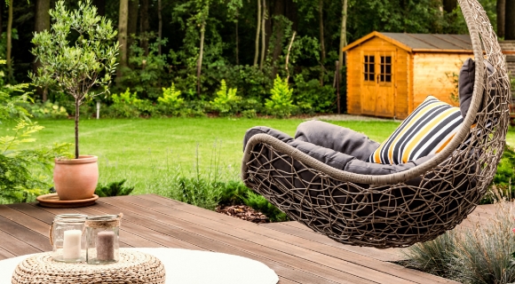 Geflochtener H&auml;ngesessel mit Kissen auf einer Terrasse aus Holz, umgeben von Gr&uuml;n und Gartenhaus.