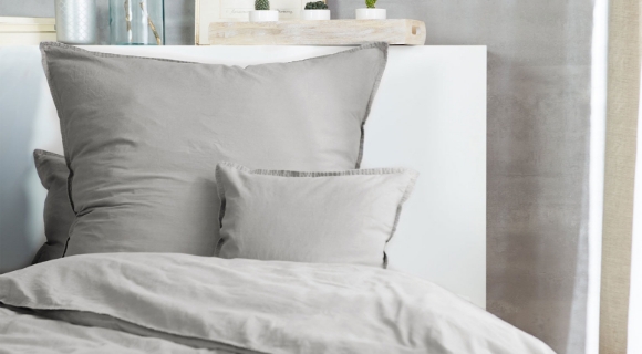Bett mit grauer Bettwäsche vor grauer Wand, dekoriert mit Pflanzen und Bildern.