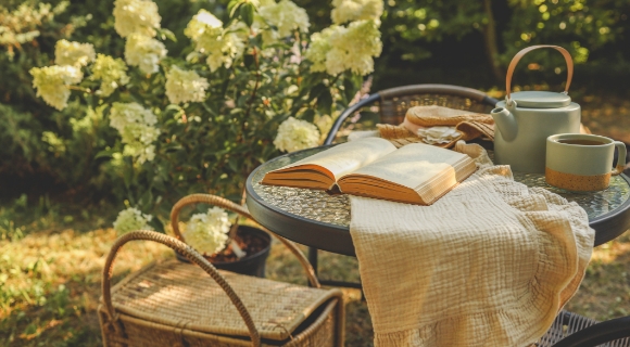 Gartentisch mit offenem Buch, Teekanne, Tasse und Stofftuch vor bl&uuml;henden Str&auml;uchern.