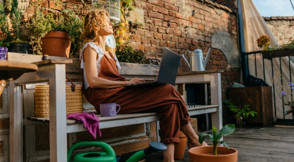 Person sitzt auf einer Bank auf einem Balkon mit einem Laptop auf dem Scho&szlig;, umgeben von warmem Sonnenlicht.
