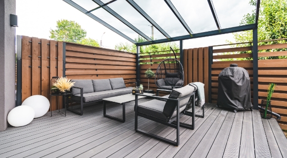 Moderne Terrasse mit &Uuml;berdachung, grauen Outdoor-M&ouml;beln, Holzverkleidung und Sitzbereich im Au&szlig;enbereich.
