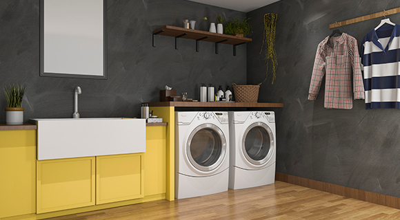 Moderner Hauswirtschaftsraum mit stilvollem Farbkontrast aus Gelb, Schwarz und Holz. Im Zentrum stehen Waschmaschine und Trockner nebeneinander unter einer Arbeitsplatte aus Holz. Links daneben befindet sich ein gelber Unterschrank mit großzügigem weißen Spülbecken. Die Rückwand ist dunkelgrau gehalten und mit einer Ablage sowie dekorativen Pflanzen und Reinigungsutensilien versehen. An der rechten Wand hängt eine Kleiderstange mit Hemd und Shirt. Der Boden ist aus hellem Holz und sorgt für eine warme Atmosphäre. Der Raum wirkt ordentlich, durchdacht und funktional – ideal für moderne Haushalte.