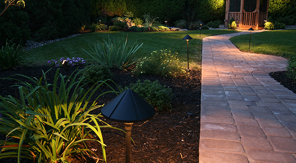 Stimmungsvoll beleuchteter Garten mit Wegleuchten, gepflegten Beeten und Holzpavillon im Hintergrund.