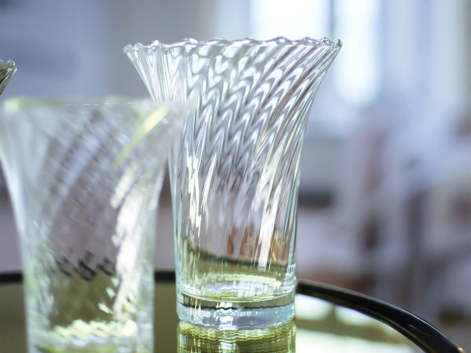 Klare Glasvase mit geschwungenem, gedrehtem Muster steht auf einer spiegelnden Oberfläche. Das Glas reflektiert das Licht und betont die elegante Struktur der Vase.